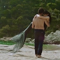 a man and woman hugging on a beach with a mermaid tail